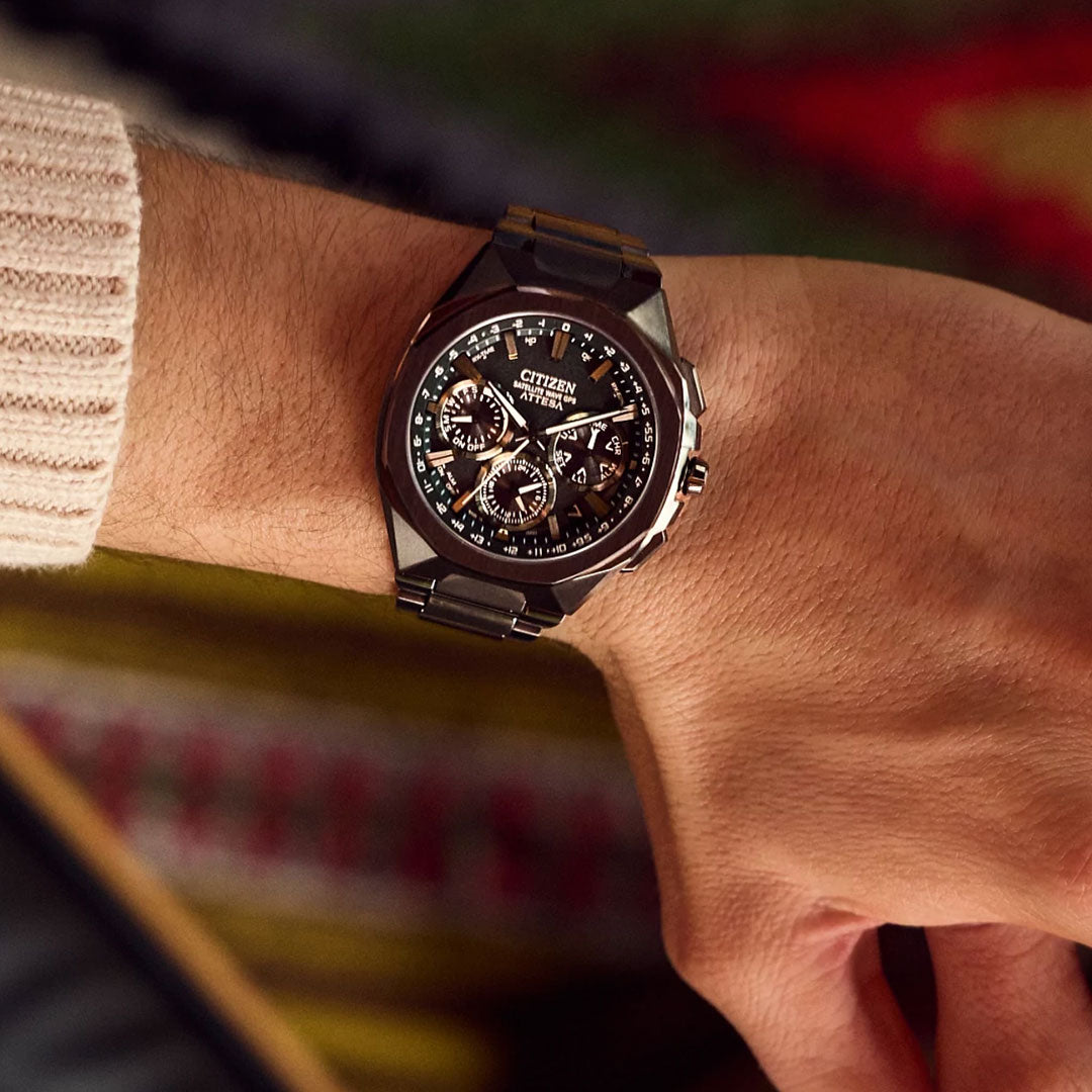 A close-up of a wrist wearing the CITIZEN Attesa Satellite Time Keeping Super Titanium Limited Edition Eco-Drive 43.2mm Watch, featuring a black band and multi-dial face against a softly blurred, warm-toned background.