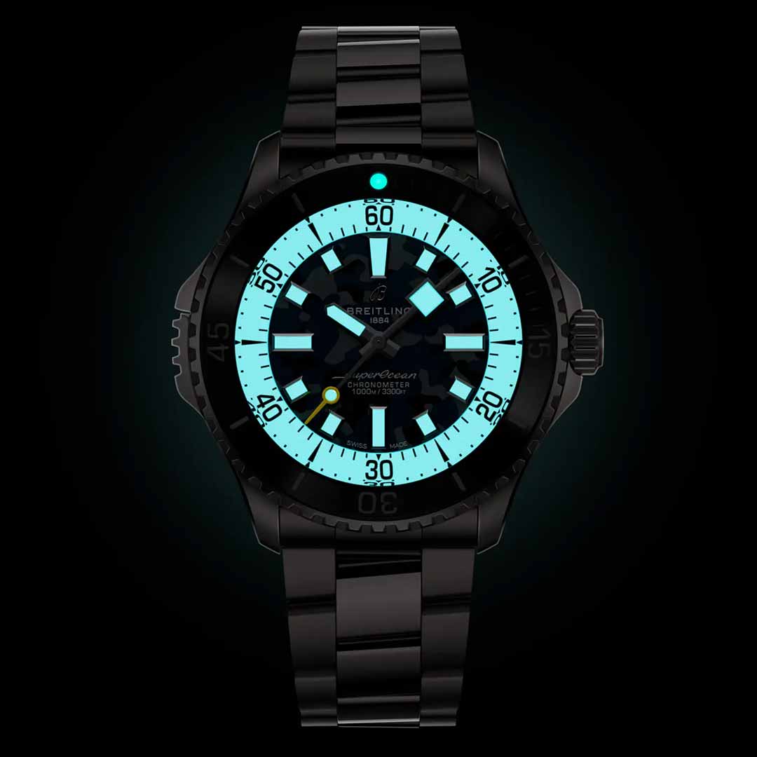 The BREITLING Superocean Automatic 46 Super Diver, by Breitling, is a dark dive watch featuring a glowing blue-green dial with luminescent indexes. It has a black metal band and showcases large numbers and hands against a black background, with the bezel marked for precise timekeeping.