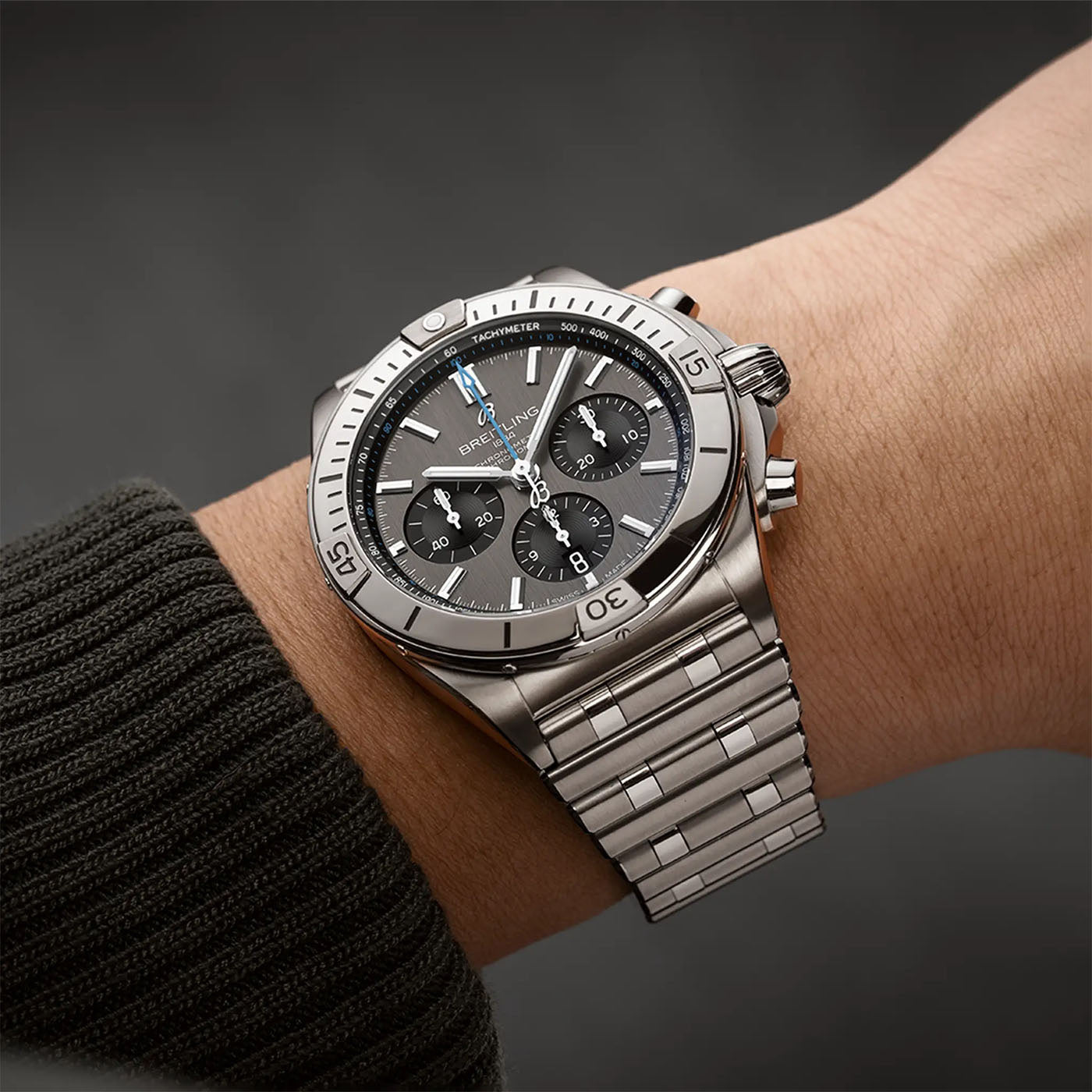 A person wearing a dark green sweater has the BREITLING Chronomat B01 42, a stainless steel watch by Breitling, on their wrist. This luxury timepiece features a black dial with silver indices, three sub-dials, and a rotating bezel, all complemented by a metal bracelet.