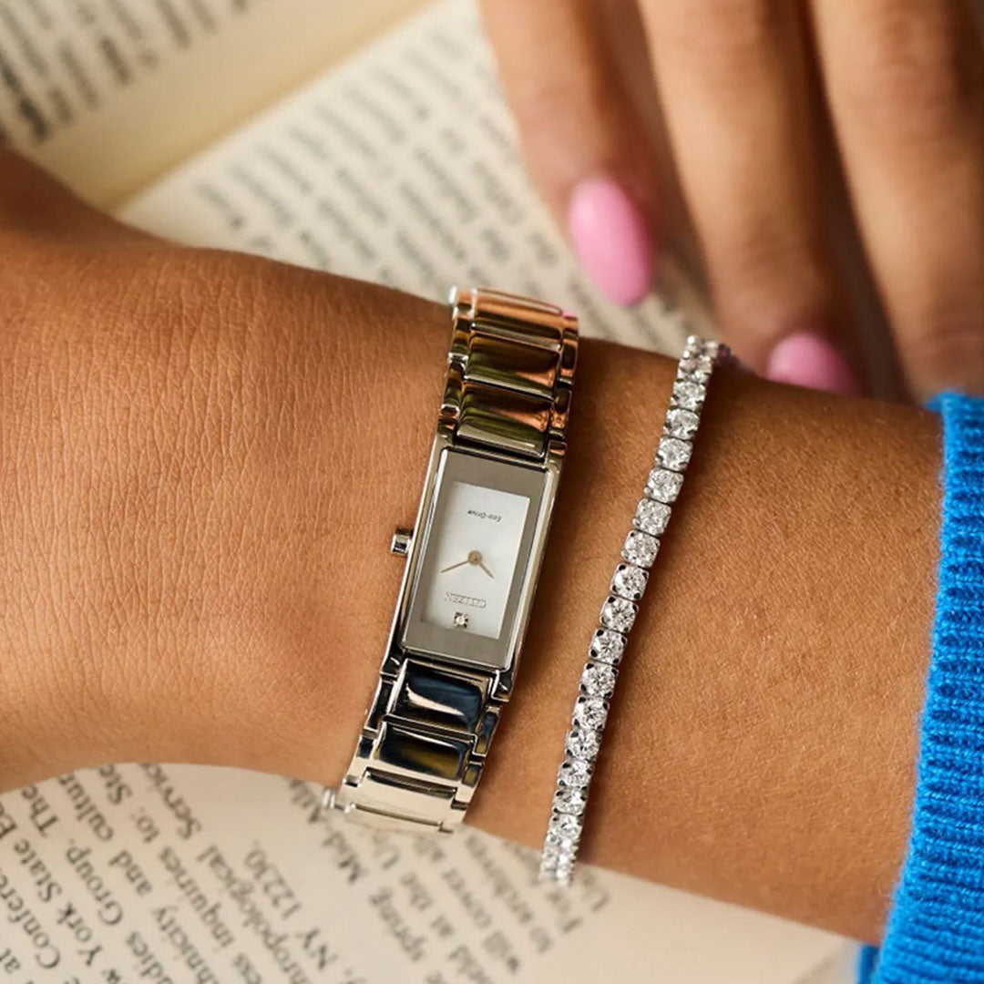 A person with pink nails wears a sleek CITIZEN Axiom Eco-Drive 14.3mm stainless steel watch and a tennis bracelet adorned with clear stones, resting their hand on an open book.