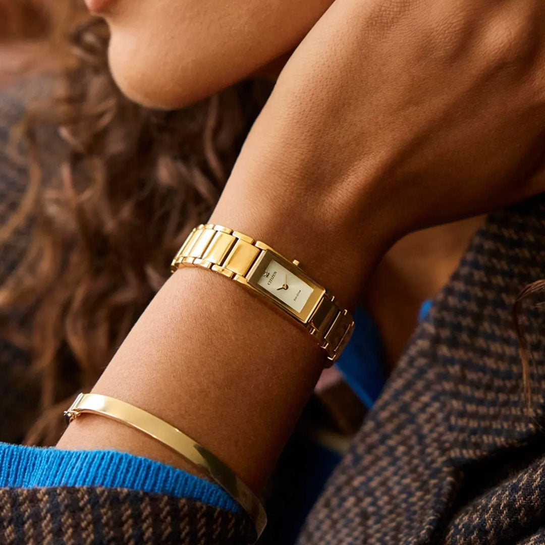 A person wearing the CITIZEN Axiom Eco-Drive 14.3mm Watch—a gold-tone dress watch with diamond markers and a gold bracelet by CITIZEN—dressed in a houndstooth-patterned coat layered over a blue sweater.