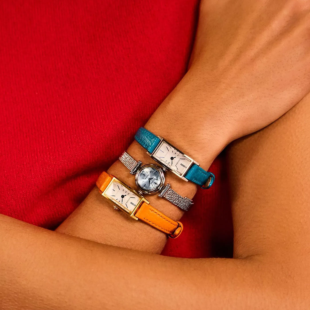 A person wearing three watches—an orange-strapped one, a blue watch, and the CITIZEN Fio Eco-Drive 18.4mm with a stainless steel mesh band—all on one arm, set against a red shirt.