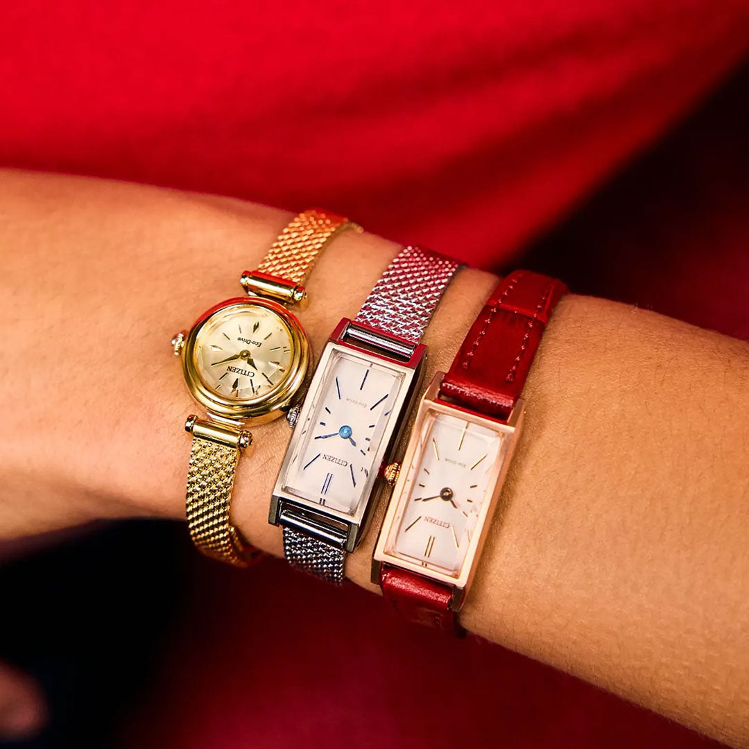 A person’s wrist wears three CITIZEN watches—a gold-tone mesh band, a rectangular silver mesh band, and the CITIZEN Fio Eco-Drive 18.4mm with a rose gold case and red leather strap—set against a bold red background.