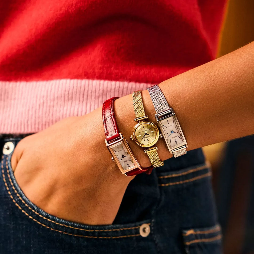 A person wears three CITIZEN watches—one with a red leather band, a gold-tone CITIZEN Fio Eco-Drive 18.4mm Watch, and another with a silver mesh band—while resting their hand in the pocket of dark blue jeans.