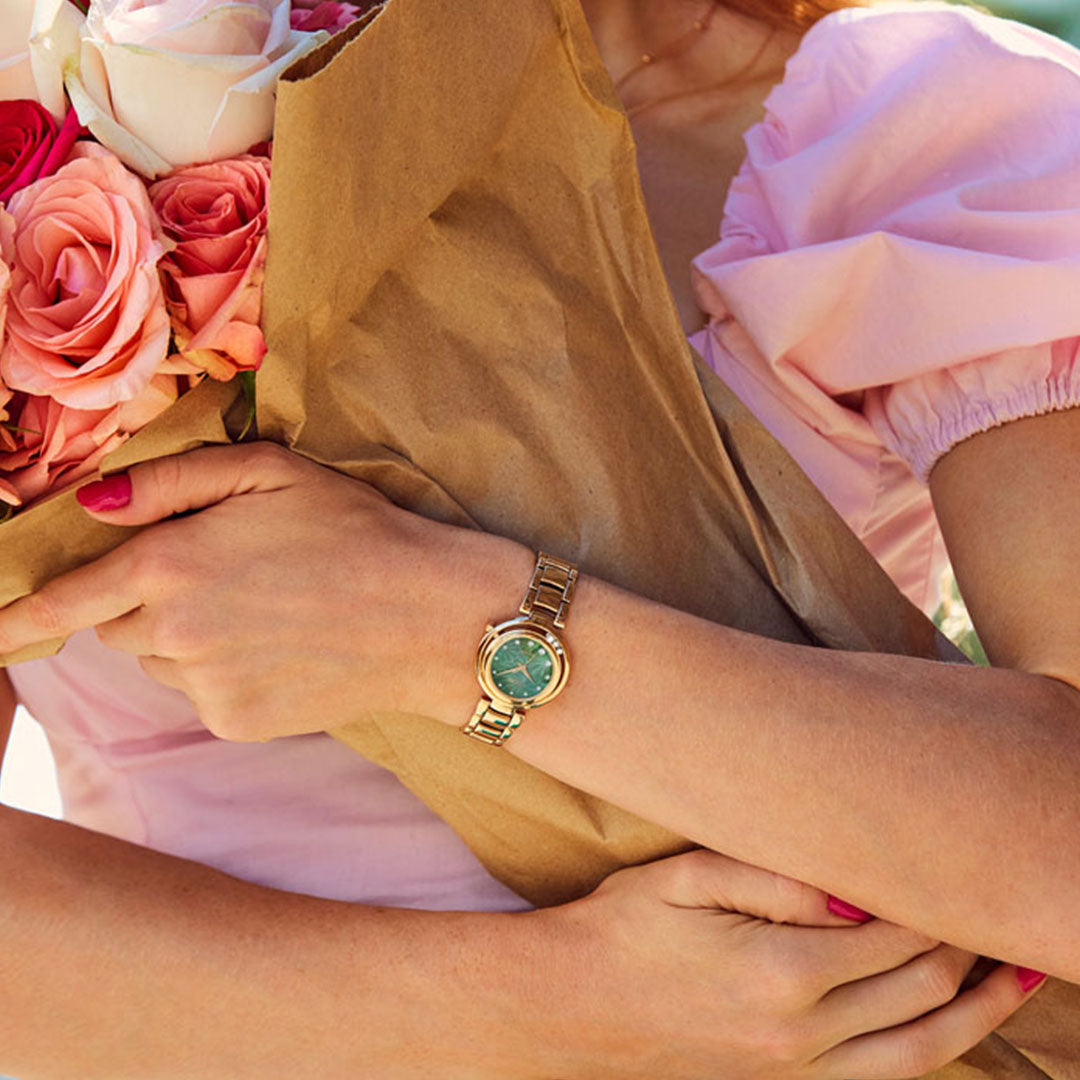 A person in a pink blouse holds a bouquet of pink and white roses wrapped in brown paper. They are wearing the CITIZEN L Arcly Eco-Drive 29.8mm Watch, featuring a sleek rose gold-tone bracelet and a green dial that catches the light beautifully.