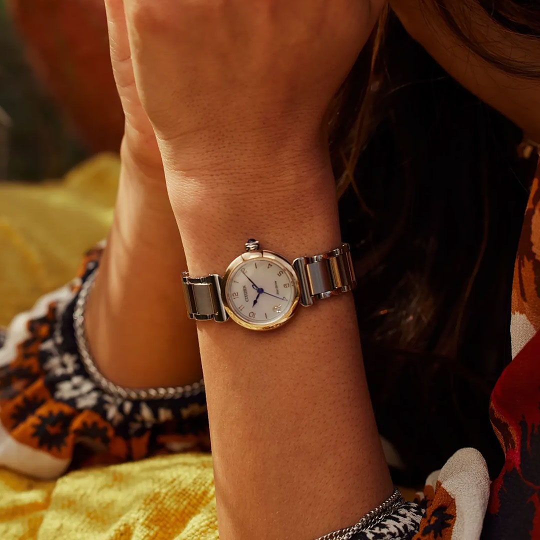 A close-up shows a CITIZEN L Mae Eco-Drive 26mm watch on a wrist, featuring a white face and blue hands. The wearer, in a colorful patterned shirt, rests their arm on a yellow cushion, reflecting CITIZEN's commitment to sustainable luxury.