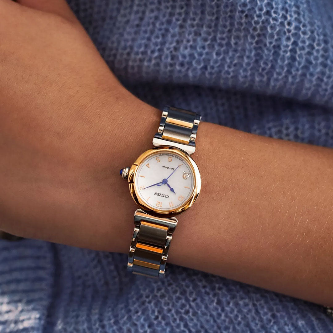A close-up shows a CITIZEN L Mae Eco-Drive 26mm Watch by CITIZEN, featuring a white round dial, gold accents, and blue hands, styled with a blue textured sweater. This sustainable luxury ladies’ timepiece combines elegance and eco-friendly design.