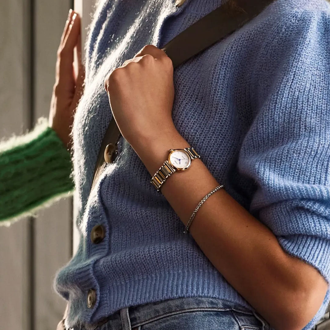 A person in a light blue knit cardigan and jeans, with a brown strap over their shoulder, showcases the CITIZEN L Mae Eco-Drive 26mm Watch by CITIZEN and a silver bracelet on their left wrist. Someone in green is nearby.