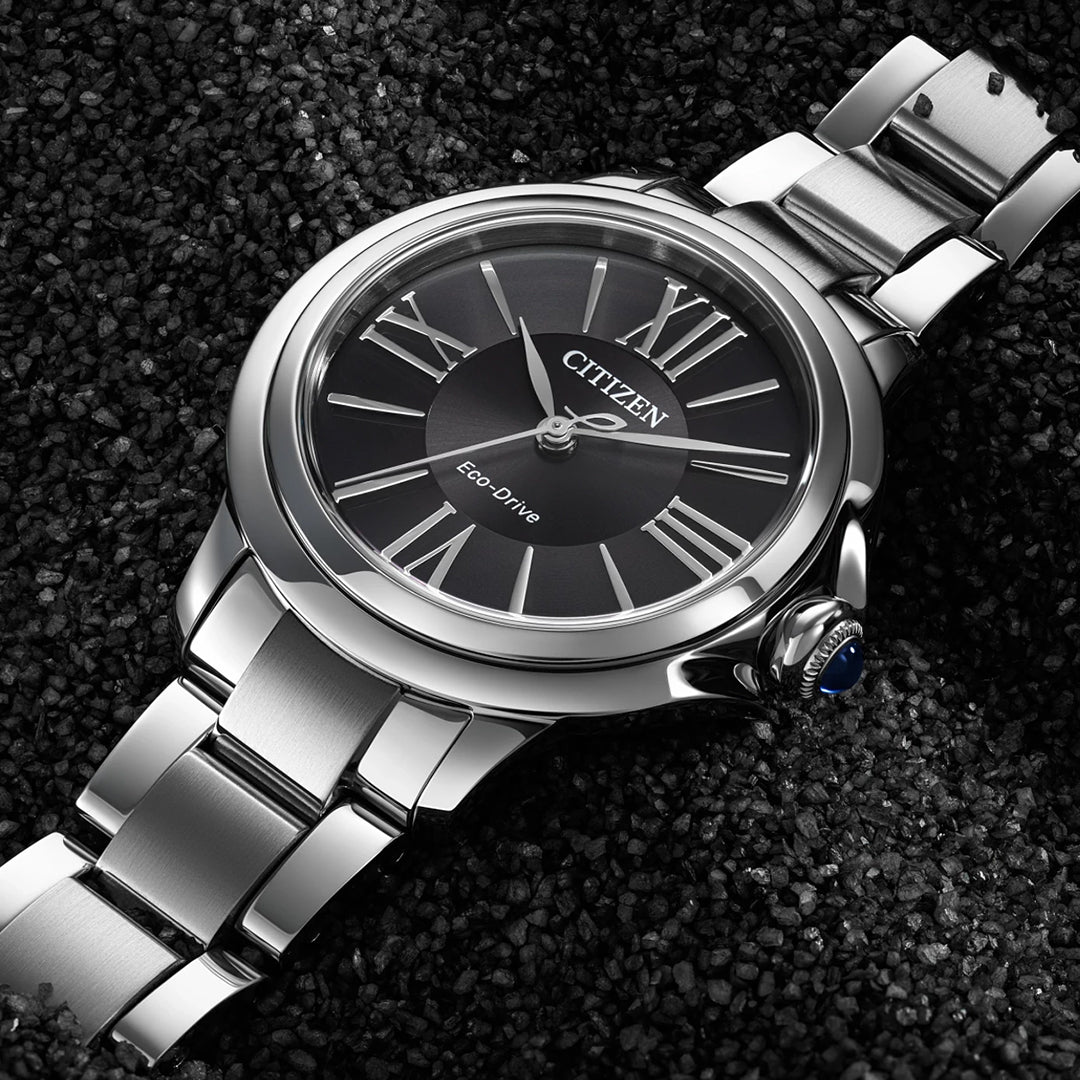 Resting on a textured black surface, the CITIZEN Le L Ceci Eco-Drive 31.5mm Watch combines elegance and sustainability, featuring a silver band, polished finish, black dial with silver Roman numerals, and a distinctive blue crown accent – a refined choice for women by CITIZEN.