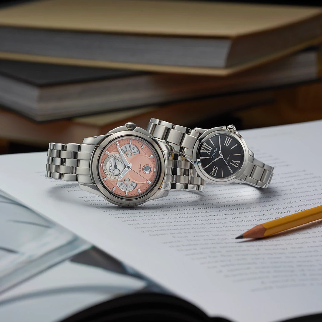 Two elegant women's watches with silver-tone stainless steel bands rest on an open book. The left is a CITIZEN Le L Ceci Eco-Drive 31.5mm Watch, boasting a pink dial with sub-dials, while the right features a black dial with Roman numerals. A nearby pencil is surrounded by books in the background.