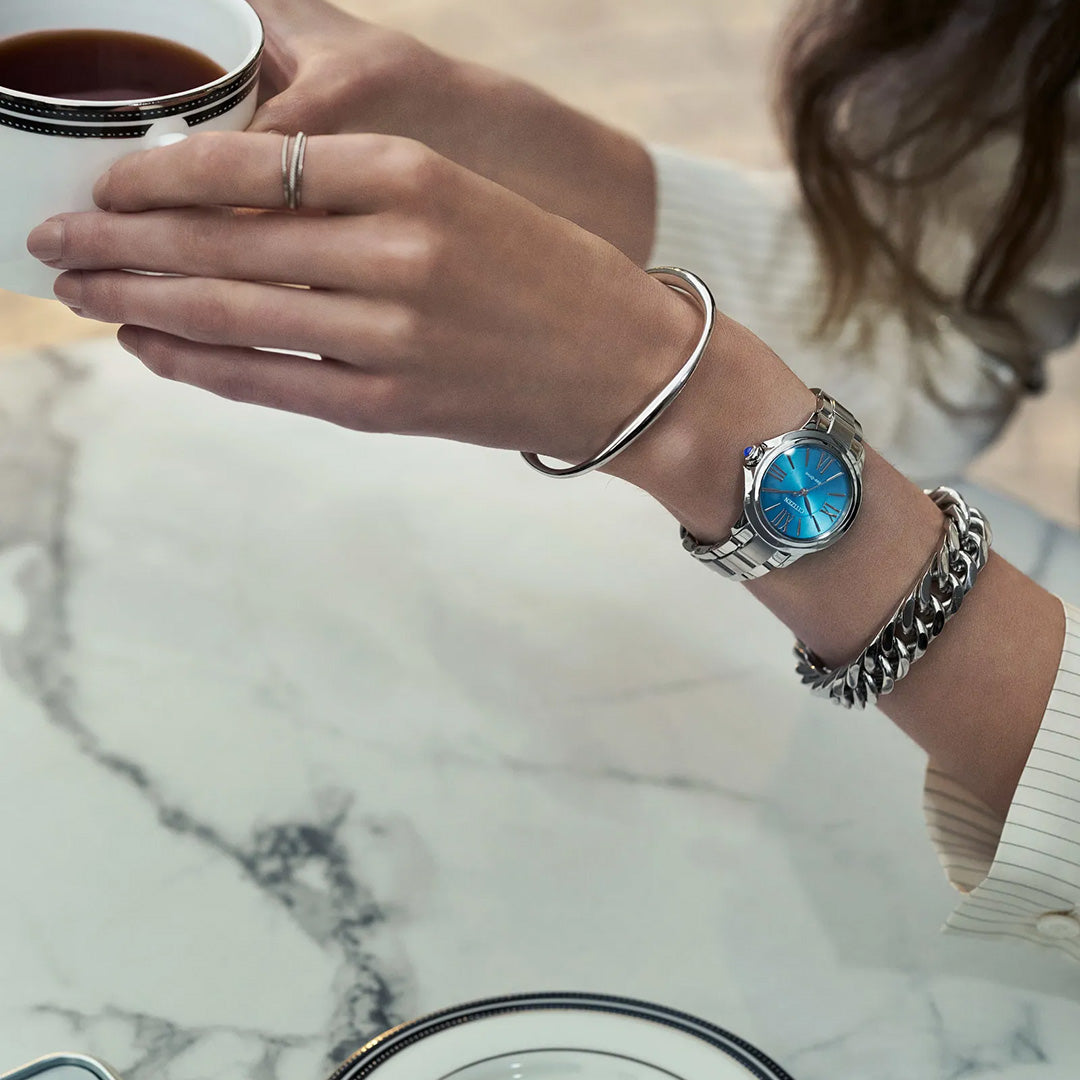 Wearing the CITIZEN Le L Ceci Eco-Drive 31.5mm Watch with its silver-tone band and blue face, a person holds a cup of coffee at a marble table, accessorized with silver bracelets and rings beside a plate.