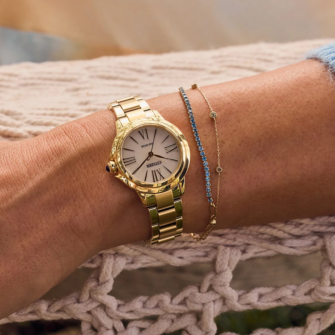 A close-up features a CITIZEN Le L Ceci Eco-Drive 31.5mm watch in gold-tone stainless steel with a white face and Roman numerals, paired with a delicate bracelet of small blue stones. This elegant scene rests on a knitted surface, exuding sophistication.