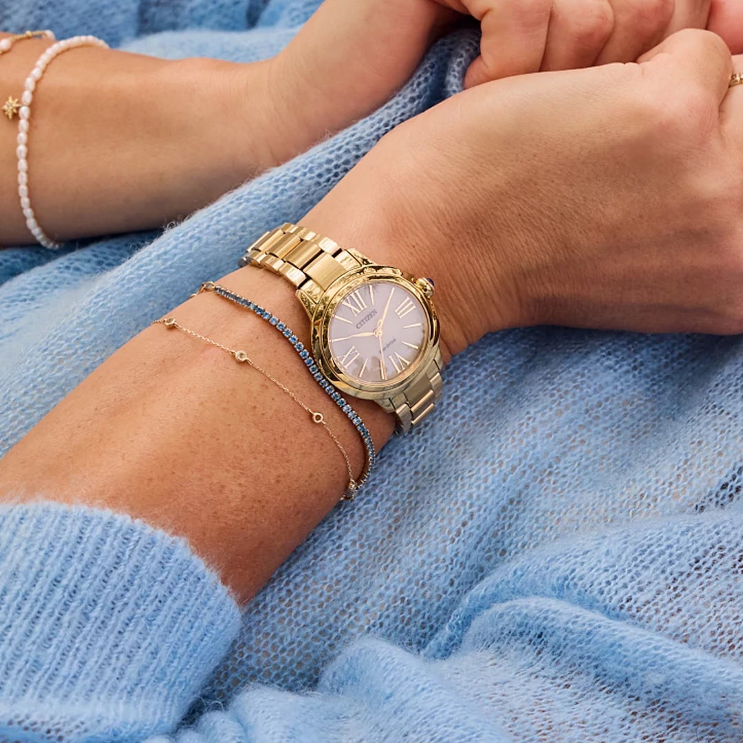 A person wearing a CITIZEN Le L Ceci Eco-Drive 31.5mm watch with an elegant gold-tone stainless steel band, Roman numeral clock face, paired with two delicate bracelets—one gold chain and one with blue beads—rests their hands gently while dressed in a soft blue sweater.