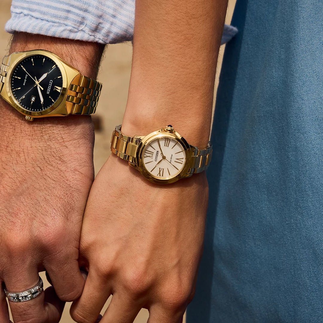 Two people holding hands wear elegant women's watches; one flaunts a gold-tone stainless steel watch with a black dial, and the other showcases a CITIZEN Le L Ceci Eco-Drive 31.5mm Watch with a white dial, all set against a beautifully blurred background.