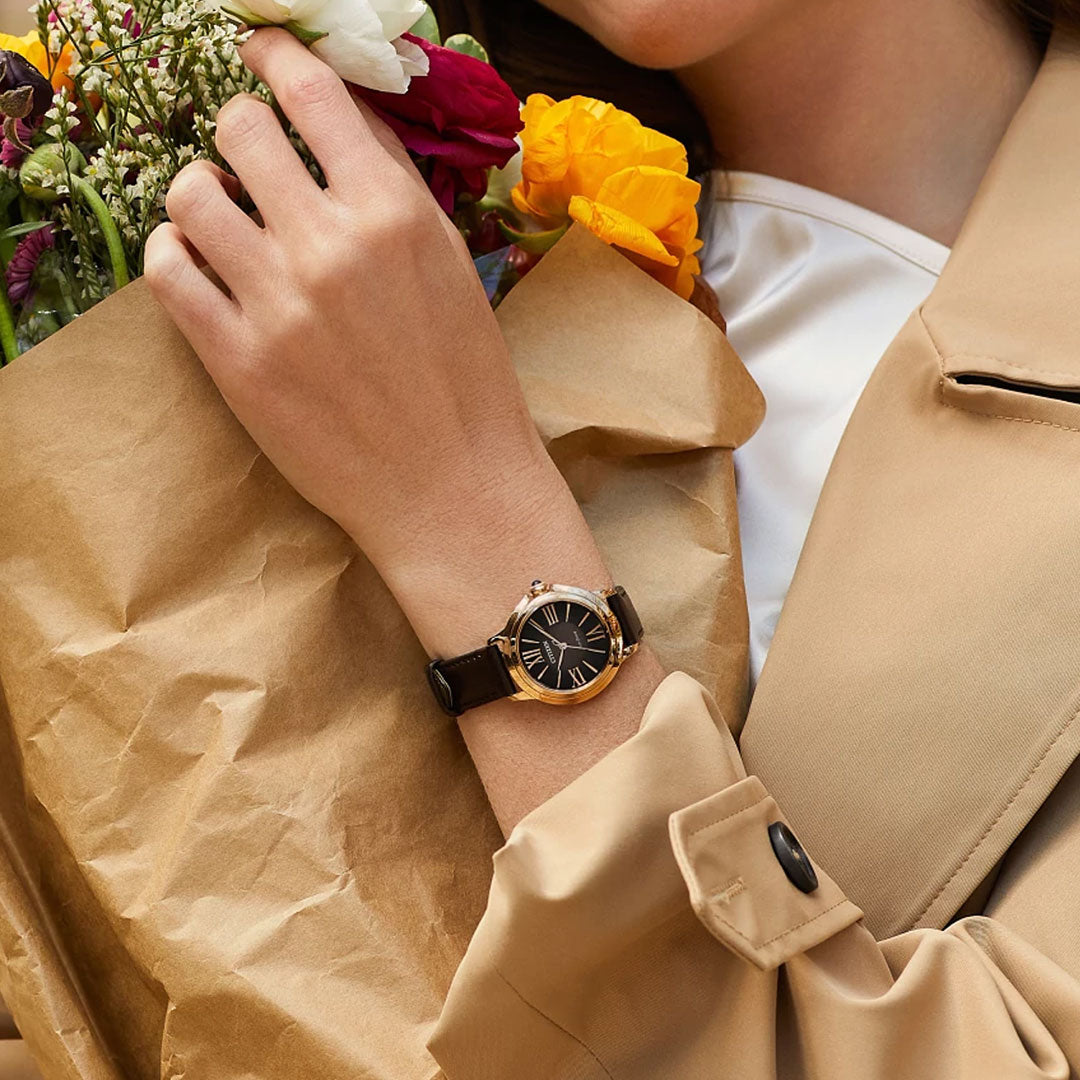 A person in a beige trench coat holds a colorful bouquet wrapped in brown paper, wearing the CITIZEN L Ceci Eco-Drive 32mm Watch by CITIZEN, featuring a black face with gold accents.