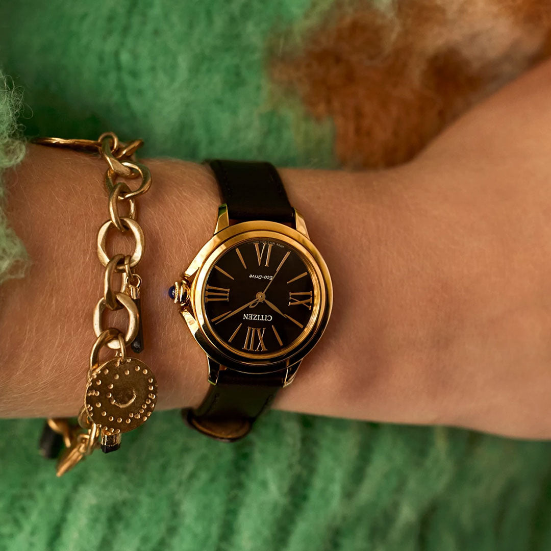 A close-up of a wrist wearing the CITIZEN L Ceci Eco-Drive 32mm Watch, featuring a gold case and black leather band, paired with a gold chain bracelet and circular charm. The person is dressed in an elegant green knit sweater.