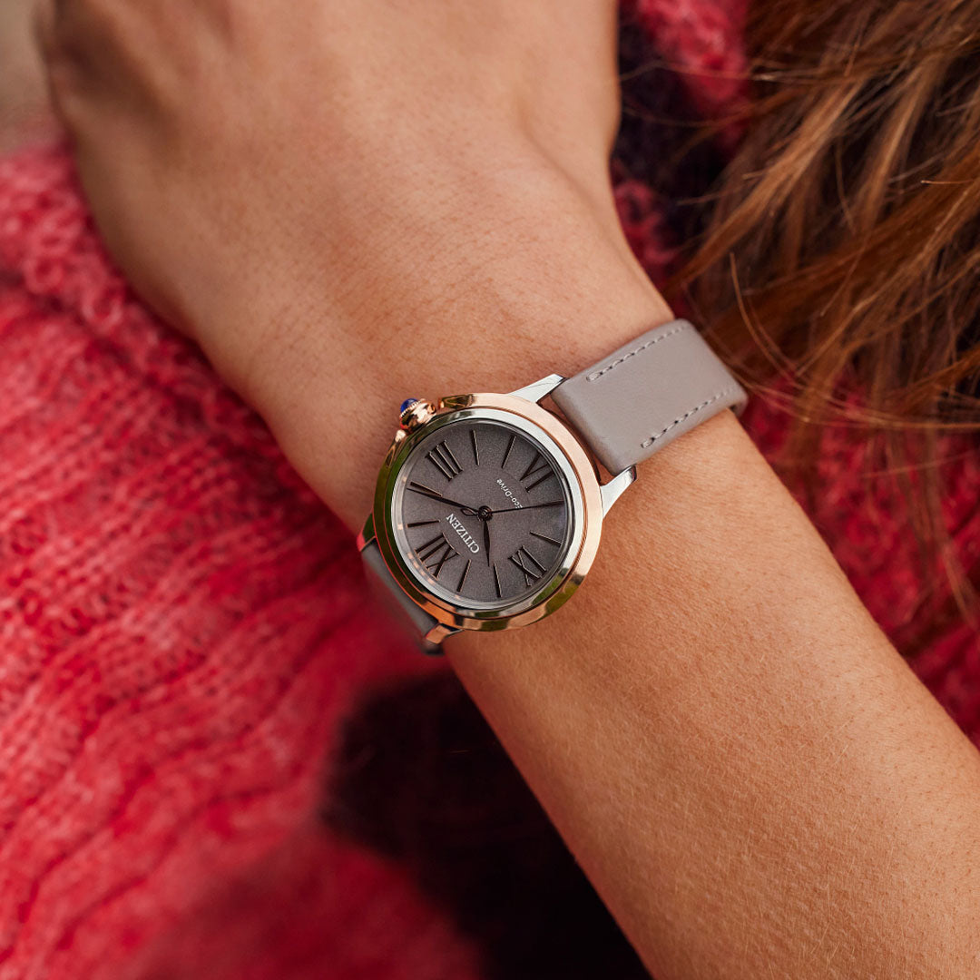 A person wearing a CITIZEN Ceci Eco-Drive 32mm watch, featuring a gray leather strap, a dark round face, and a gold-colored bezel. The elegant timepiece beautifully complements their textured red sweater.