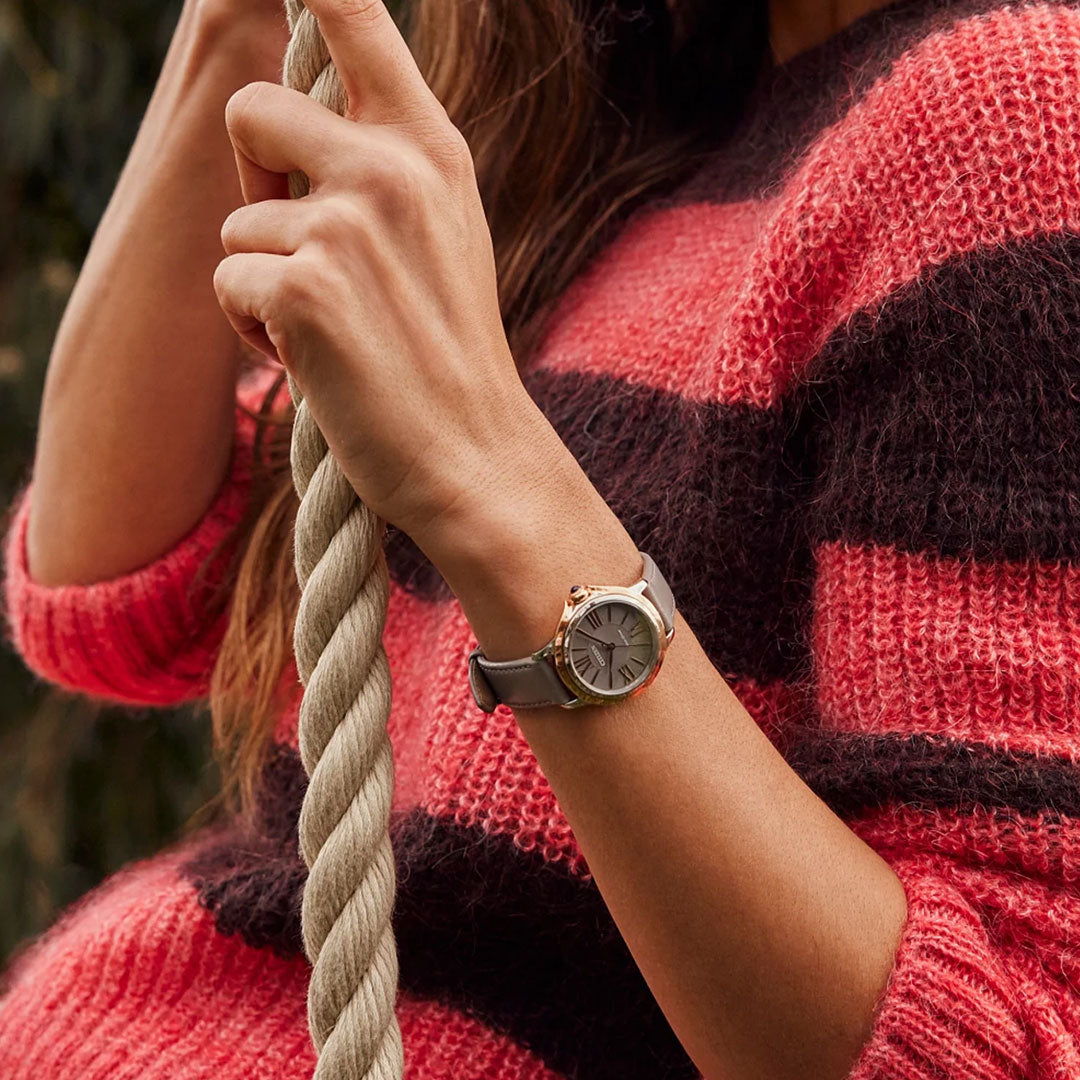 A person wearing a red and black striped sweater is holding a thick rope. On their left wrist, they wear the CITIZEN Ceci Eco-Drive 32mm Watch, featuring an elegant round face and leather strap that evokes the classic style of a Citizen dress watch. The background is blurred, highlighting the details of the watch and sweater.