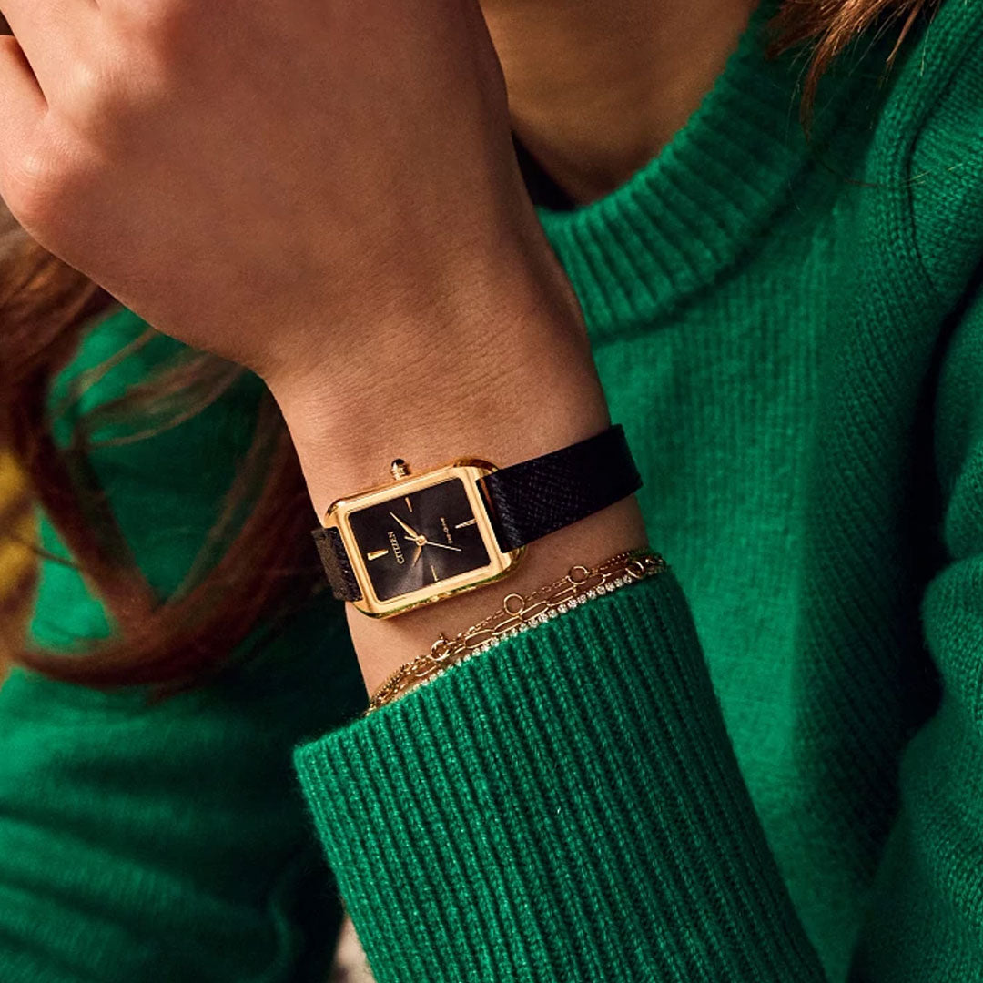 A person in a green knit sweater wears the CITIZEN Dress Classic Eco-Drive 23.5mm Watch, featuring a yellow gold-tone stainless steel case, minimalist dial, black strap, and a delicate gold bracelet.
