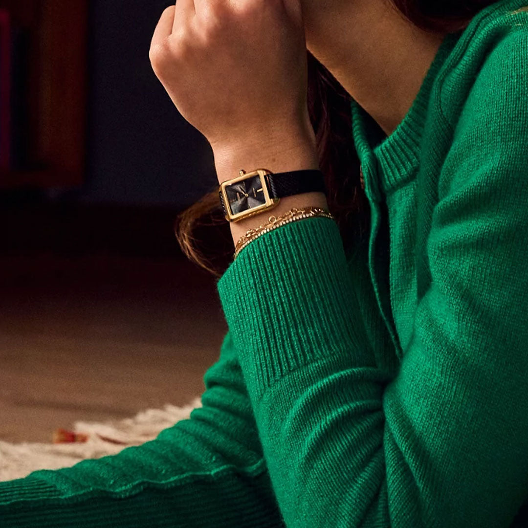 A person in a green sweater rests their chin on their hand, displaying the CITIZEN Dress Classic Eco-Drive 23.5mm Watch with a minimalist black and gold dial and a thin chain bracelet on their wrist.