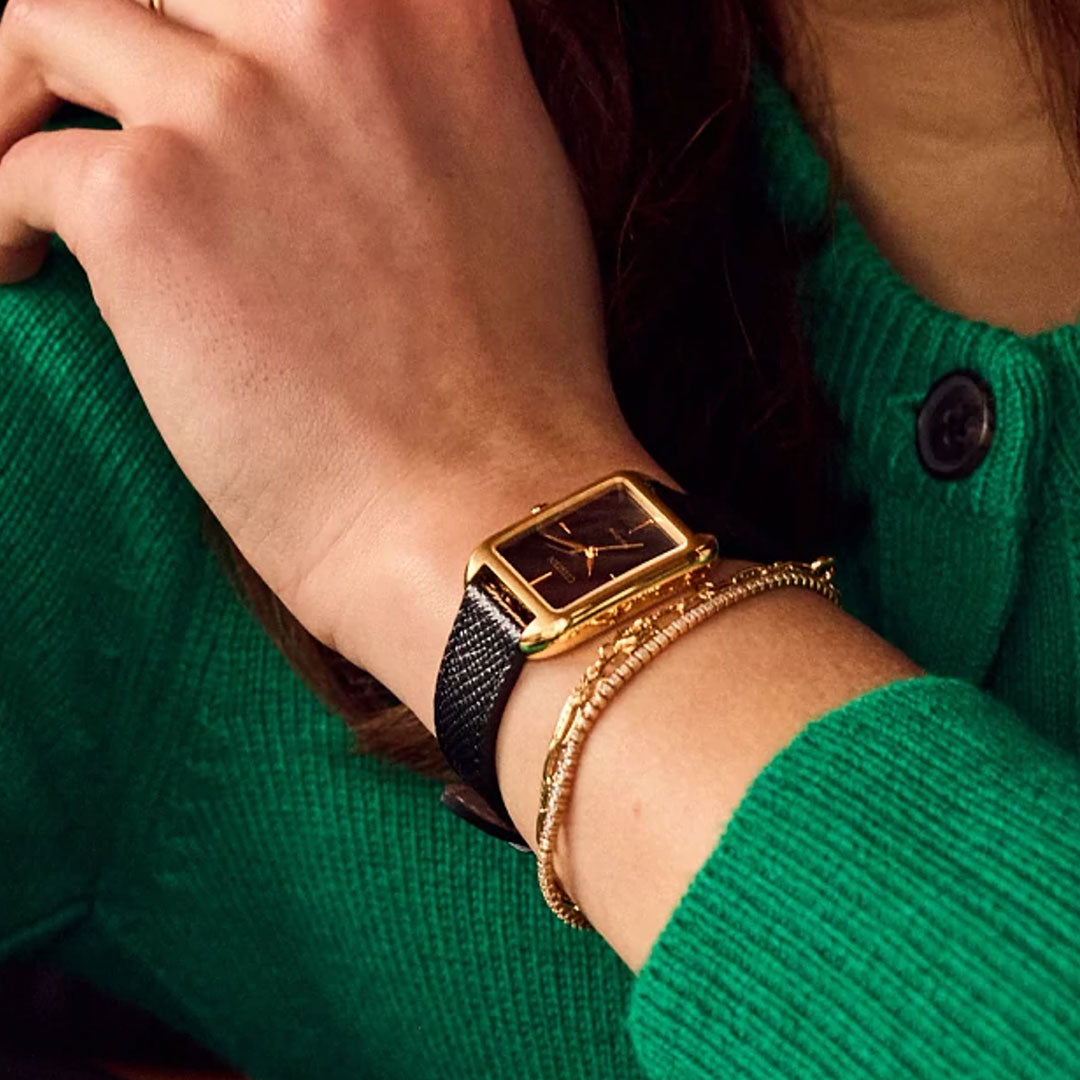 Wearing a green sweater, a person rests their chin on their hand, showcasing the CITIZEN Dress Classic Eco-Drive 23.5mm Watch with a minimalist dial and a delicate gold bracelet on their wrist.