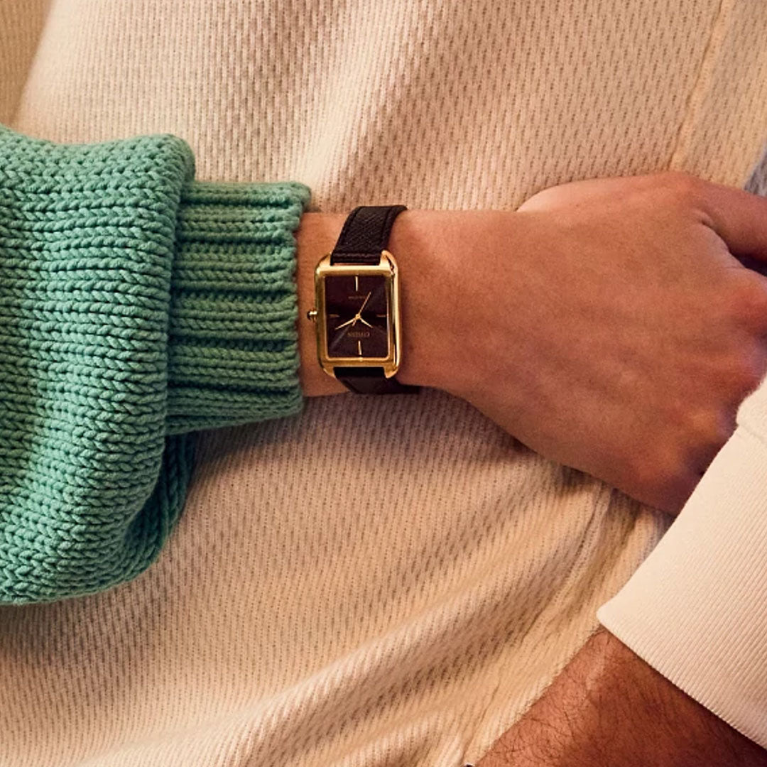 A close-up of a person wearing the CITIZEN Dress Classic Eco-Drive 23.5mm Watch with a gold-tone stainless steel case, black leather strap, and minimalist dial, styled with a cream textured sweater and another arm in a green knit sleeve.