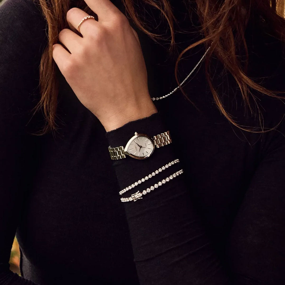 A woman wears a black long-sleeve top, the CITIZEN L Rainell Eco-Drive 26mm Watch by CITIZEN in silver-tone stainless steel, two silver bracelets, a thin silver necklace, and a gold ring. Her hand is near her neck with brown hair visible.
