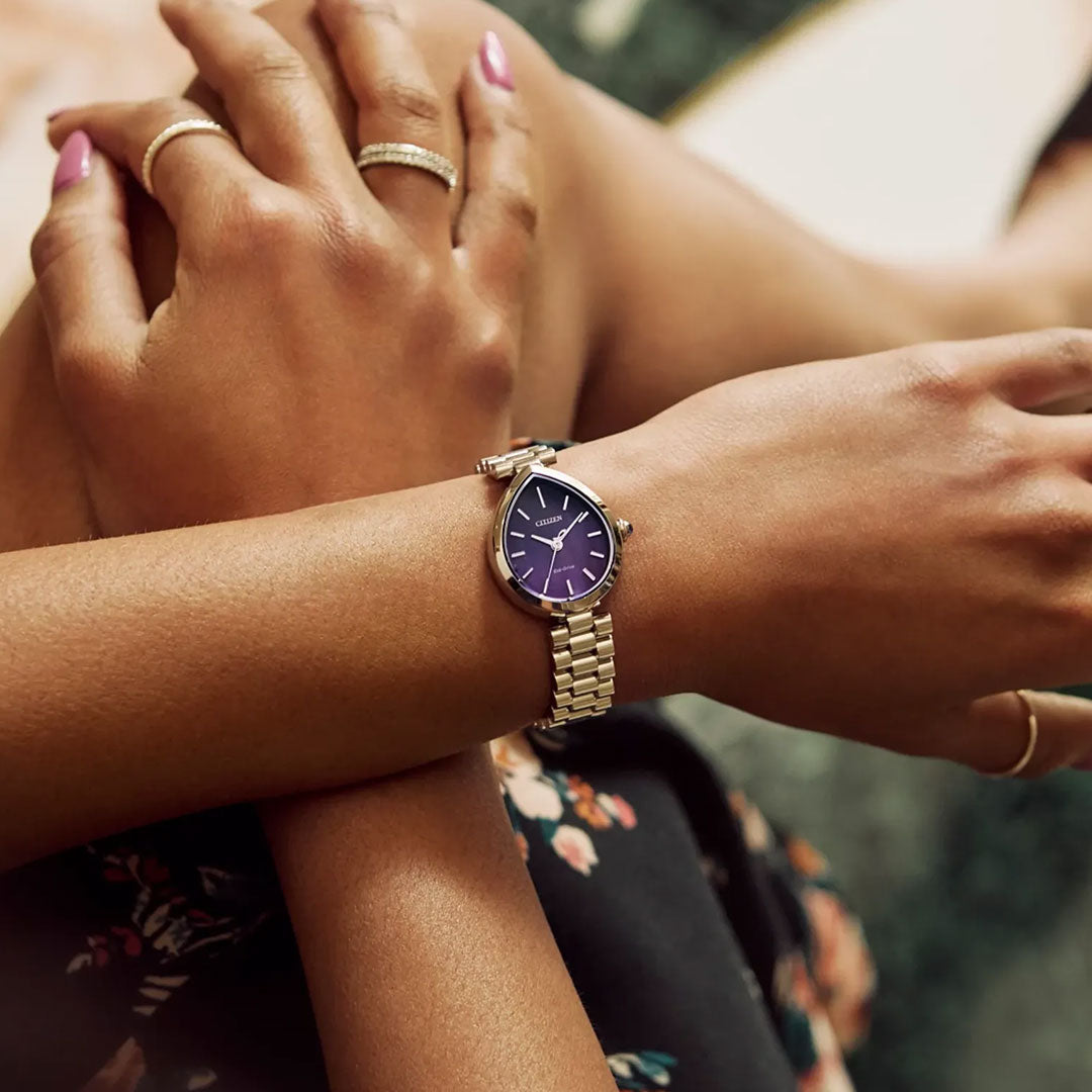 A close-up shows crossed arms wearing the CITIZEN L Rainell Eco-Drive 26mm Watch by CITIZEN, featuring a purple face and stainless steel band, with neatly manicured hands wearing a gold ring against a floral outfit.