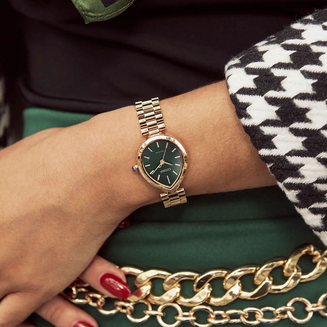 A woman’s wrist features the CITIZEN L Rainell Eco-Drive 26mm watch by CITIZEN, with a rose gold-tone and green dial. She wears a black-and-white houndstooth jacket, green skirt, gold chain belt, red nails, and a patterned scarf.