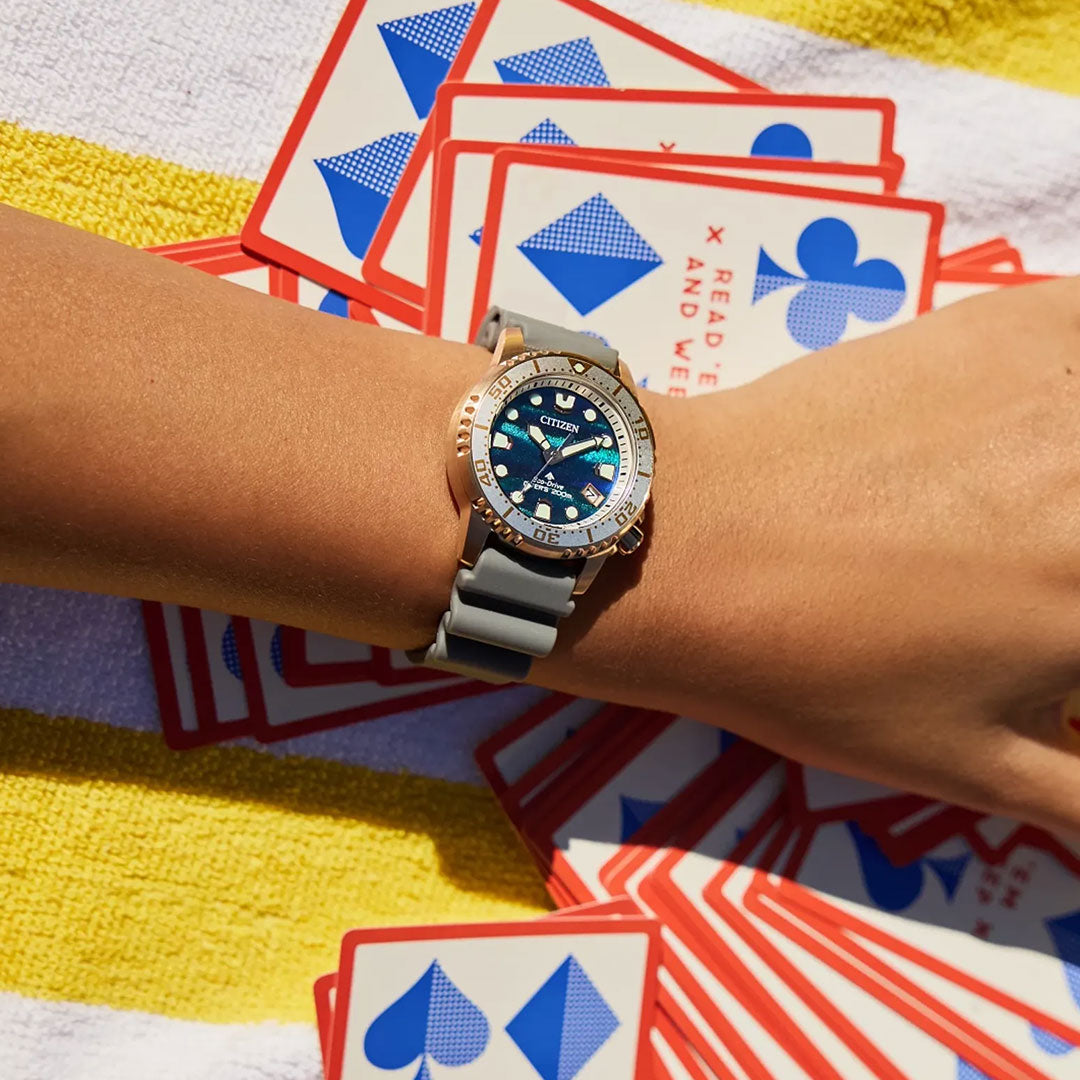 Someone wears the CITIZEN Promaster Dive Eco-Drive 36.5mm Watch Limited Edition with a grey strap, resting their arm on a yellow and white towel amidst scattered playing cards.