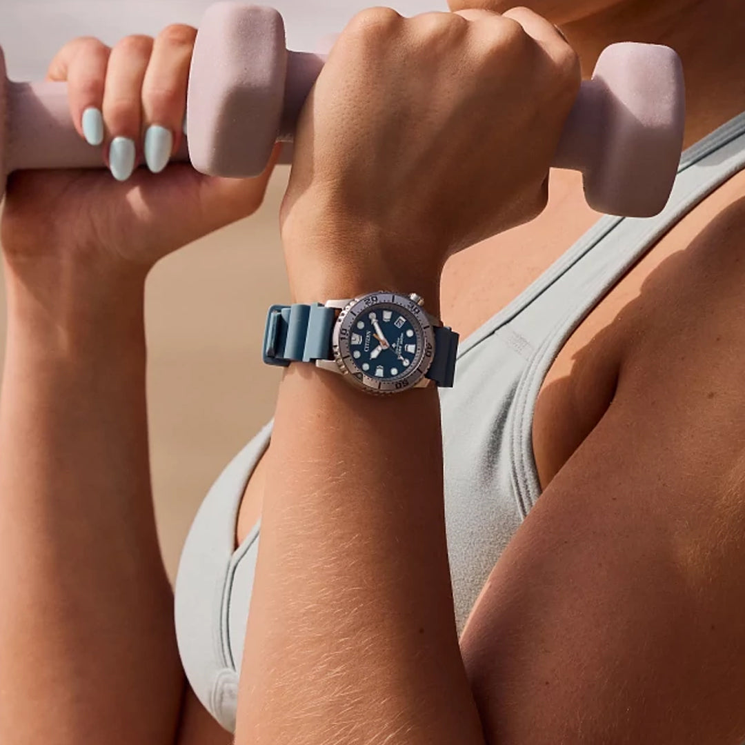 A person in a light gray sports bra holds pink dumbbells near their chest, showcasing the CITIZEN Promaster Dive Eco-Drive 37mm Watch. This sleek timepiece features a large silver bezel and blue strap, utilizing Eco-Drive technology. The background is blurred.