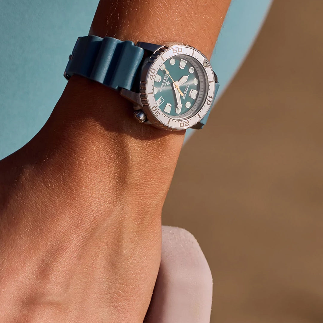 A close-up showcases a wrist with the CITIZEN Promaster Dive Eco-Drive 37mm watch, featuring a blue theme, silver accents, and Eco-Drive tech. A soft fabric is held in the other hand against a blurred brown and teal backdrop enhancing its elegance.