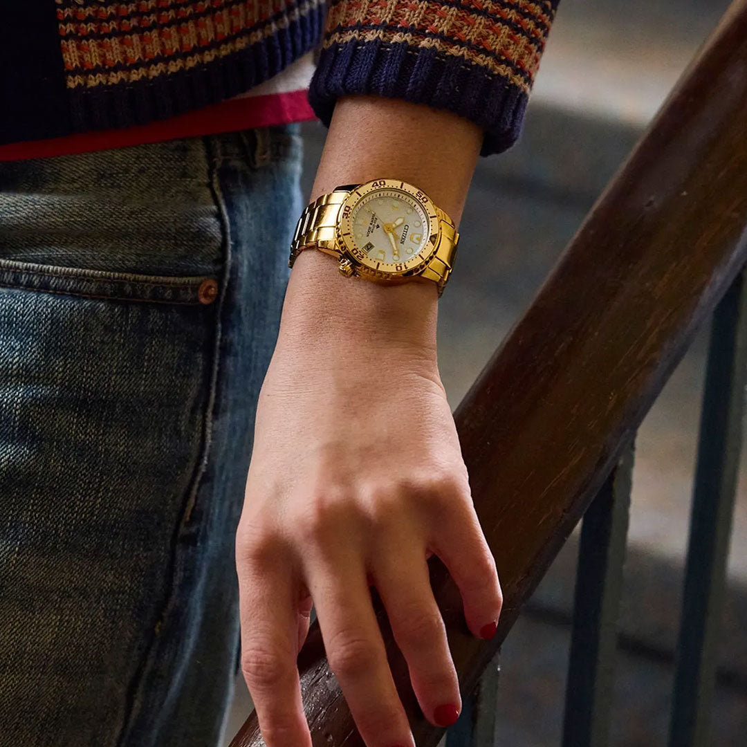 Wearing blue jeans, a colorful knit sweater, and red nail polish, a person rests their hand on a wooden railing, showcasing the CITIZEN Promaster Dive Eco-Drive 36.5mm Watch by CITIZEN.