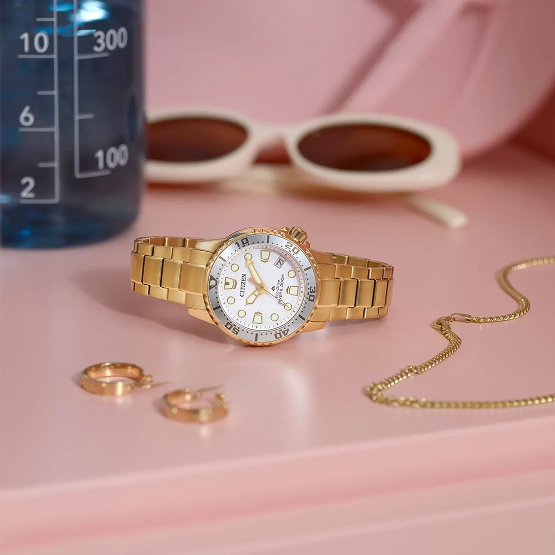 A CITIZEN Promaster Dive Eco-Drive 36.5mm Watch with a white face is displayed on a pink surface beside a gold necklace, gold hoop earrings, white-framed sunglasses, and a blue beaker.