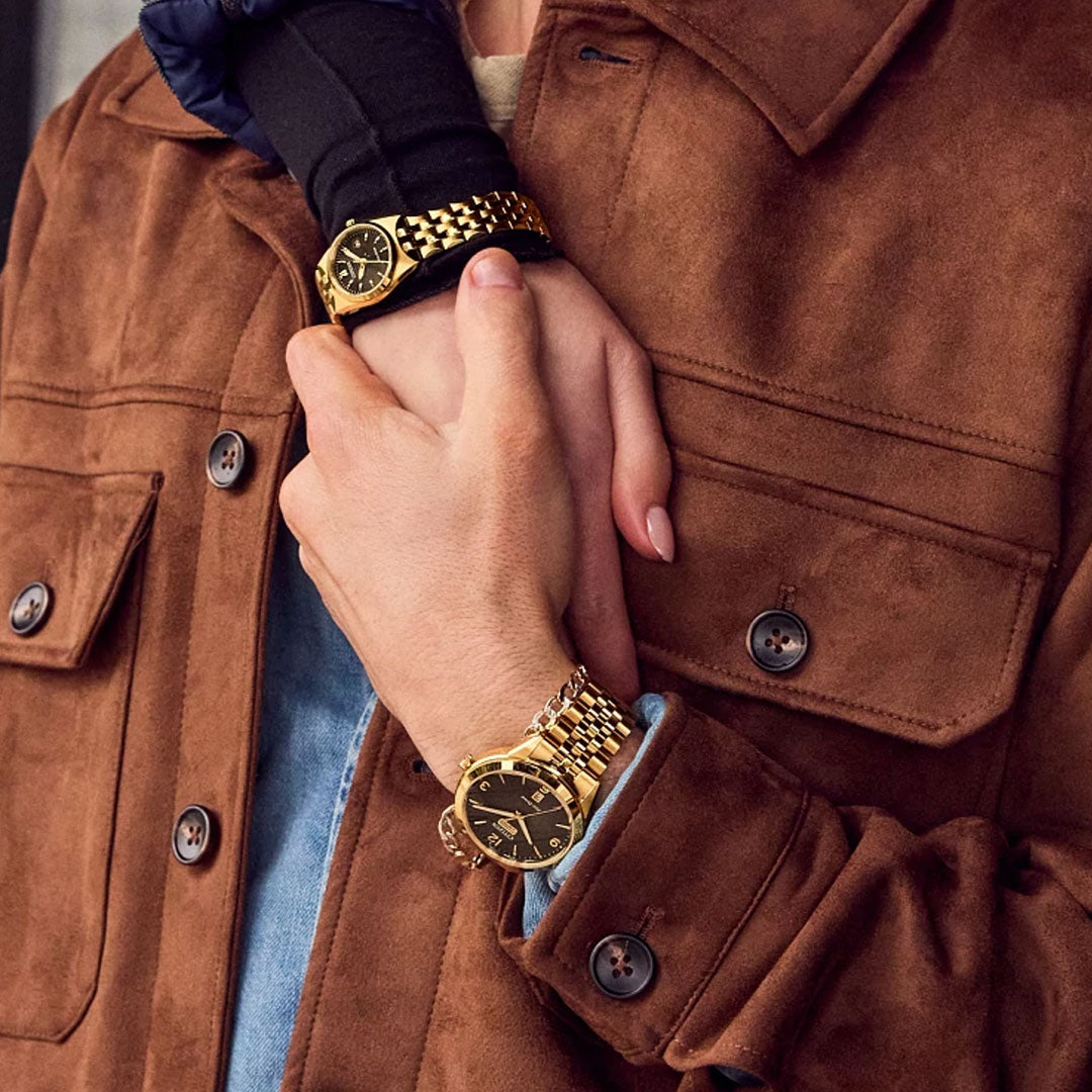Two people in brown suede jackets and gold watches gently hold hands; one wears a CITIZEN Corso Eco-Drive 27mm Watch, both featuring black faces and gold-tone stainless steel bands.