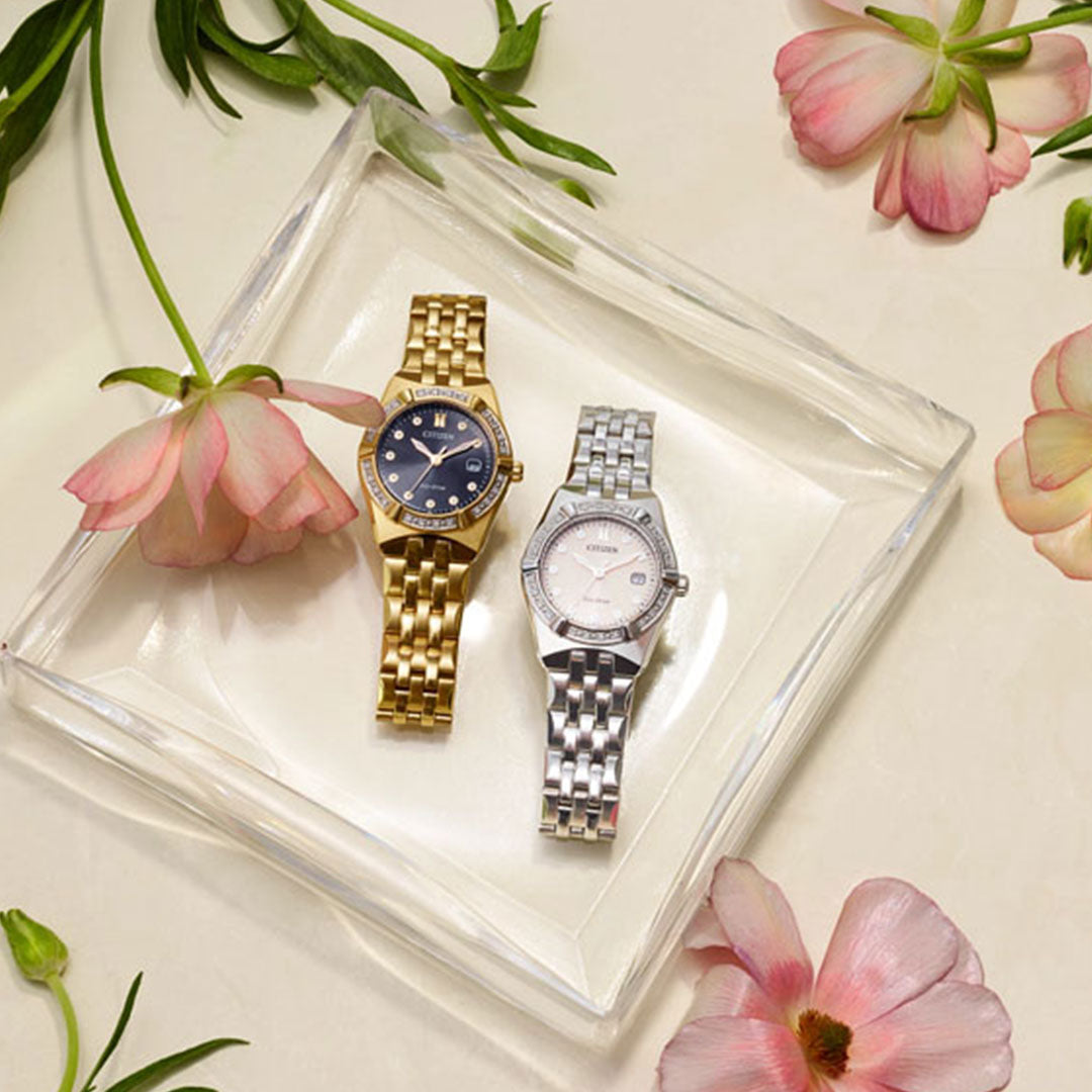 Two sophisticated ladies' watches from CITIZEN are showcased on a transparent square tray surrounded by pink flowers. One watch, the CITIZEN Corso Diamond Eco-Drive 28mm, features Eco-Drive technology along with a gold band and black dial; the other flaunts a silver band with a diamond-accented bezel and white dial. The light-colored background is elegantly decorated with scattered green leaves.