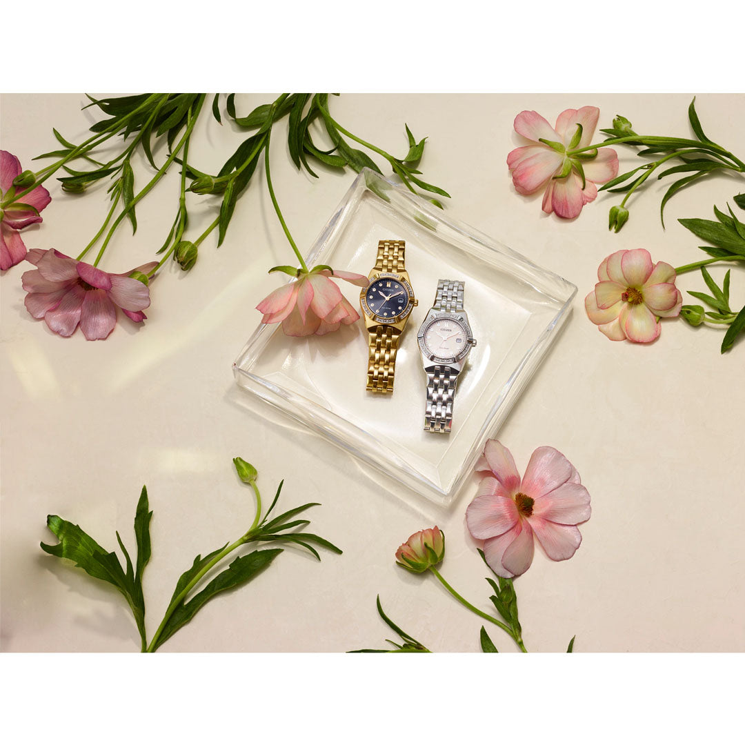 On a light background, two elegant CITIZEN ladies' wristwatches rest on a clear tray surrounded by pink flowers and green stems. One watch, the CITIZEN Corso Diamond Eco-Drive 28mm, showcases a gold band with a dark face and a diamond-accented bezel. The other gleams with its silver band and light face.