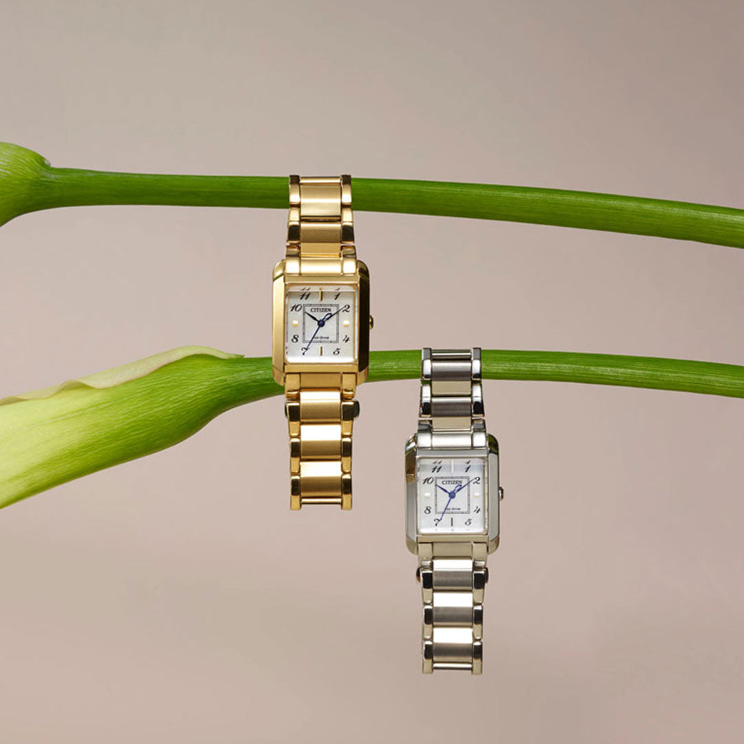 Two elegant wristwatches with square dials hang on a green stem. One is a gold CITIZEN Bianca, while the other is a stainless-steel CITIZEN Bianca Eco-Drive 21.5mm Watch. The background is a muted, light beige, enhancing the watches' metallic sheen.