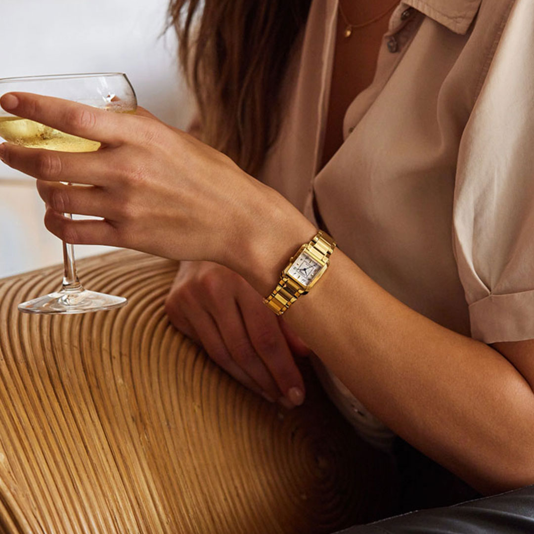 A person in a beige shirt holding a glass of white wine is resting their arm on a wicker chair. They are wearing the CITIZEN Bianca Eco-Drive 21.5mm Watch with a square face on their left wrist, crafted from gold-tone stainless steel.