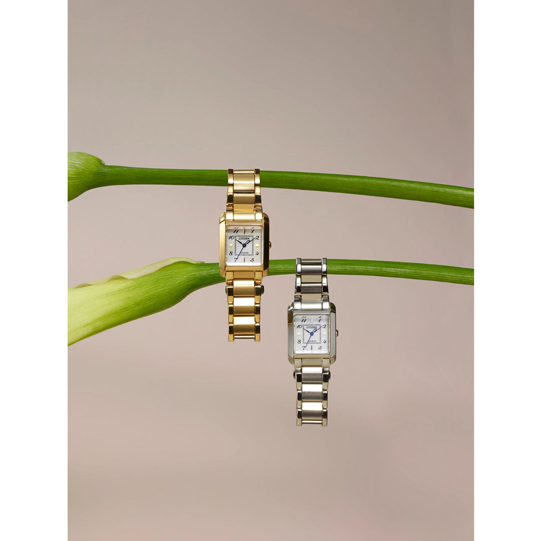 Two CITIZEN Bianca Eco-Drive 21.5mm watches with metal link bands, one in gold-tone stainless steel and the other in silver, are elegantly draped over a green stem with white flowers. These CITIZEN timepieces feature square faces that precisely display the time, set against a soft, neutral backdrop.