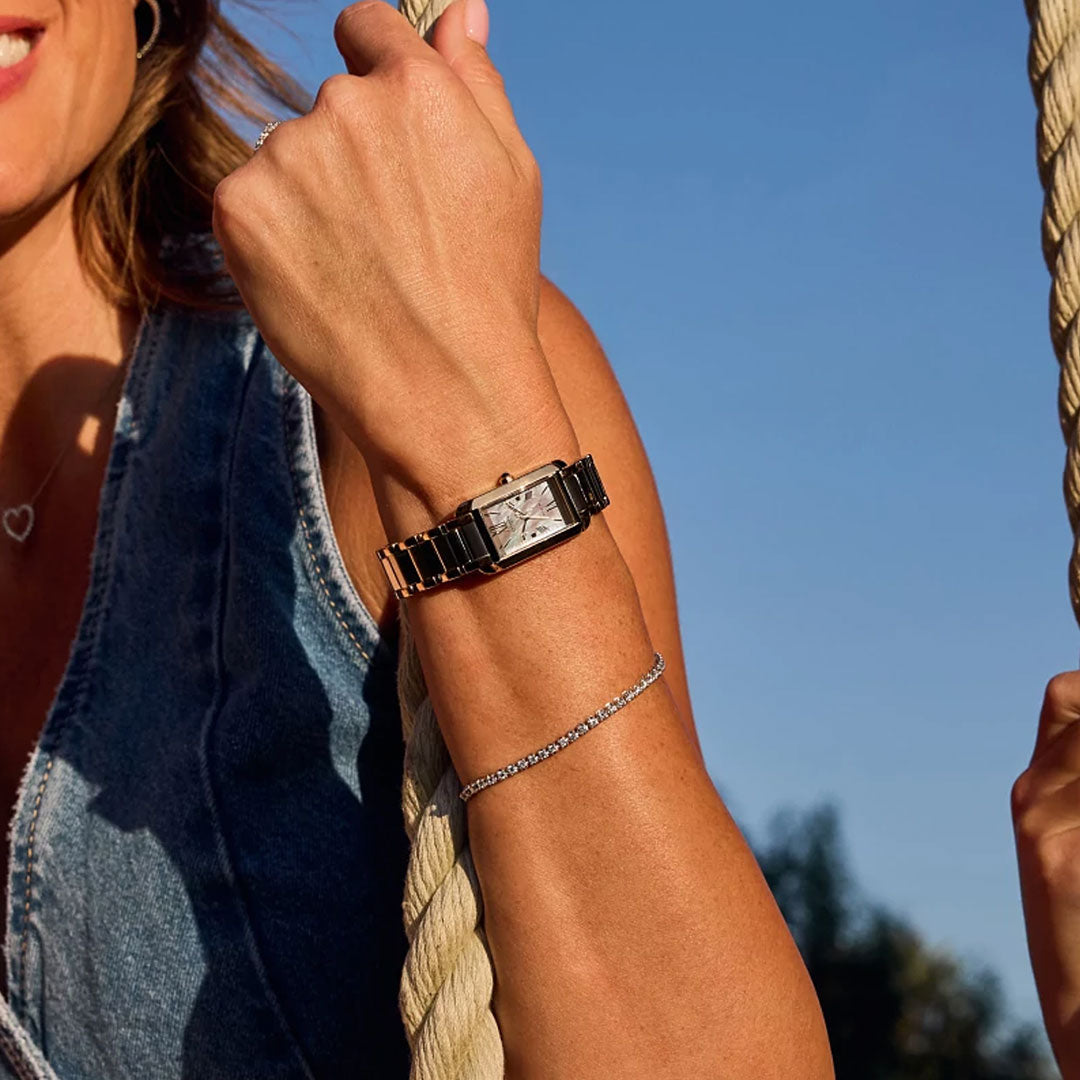 Outdoors under a clear blue sky, a person in a sleeveless denim top holds a rope, showcasing the CITIZEN L Bianca Eco-Drive 19.5mm Watch and a delicate chain bracelet.