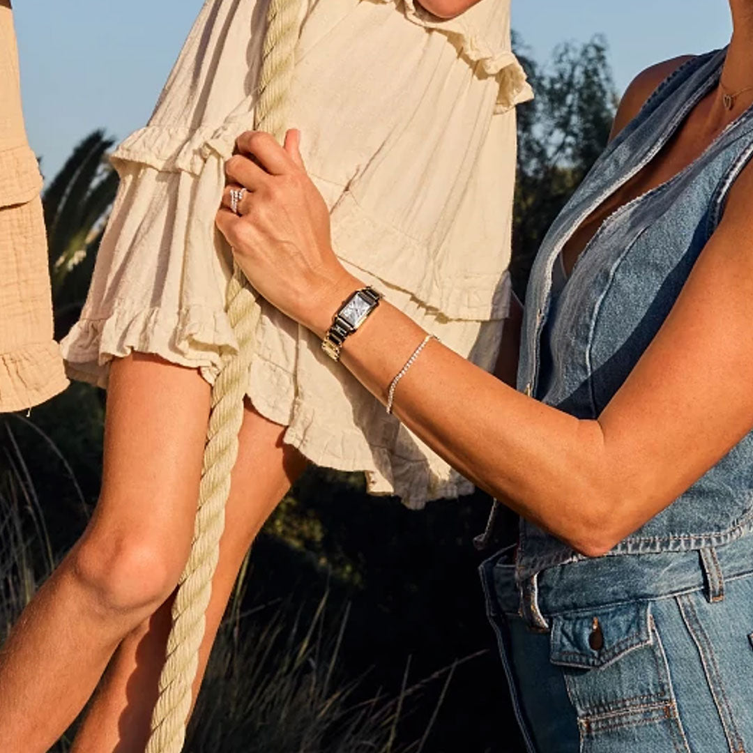 Wearing the CITIZEN L Bianca Eco-Drive 19.5mm Watch, a woman in a sleeveless denim dress stands by a rope swing outdoors, embodying sustainable luxury from CITIZEN amid lush greenery with timeless, effortless style.