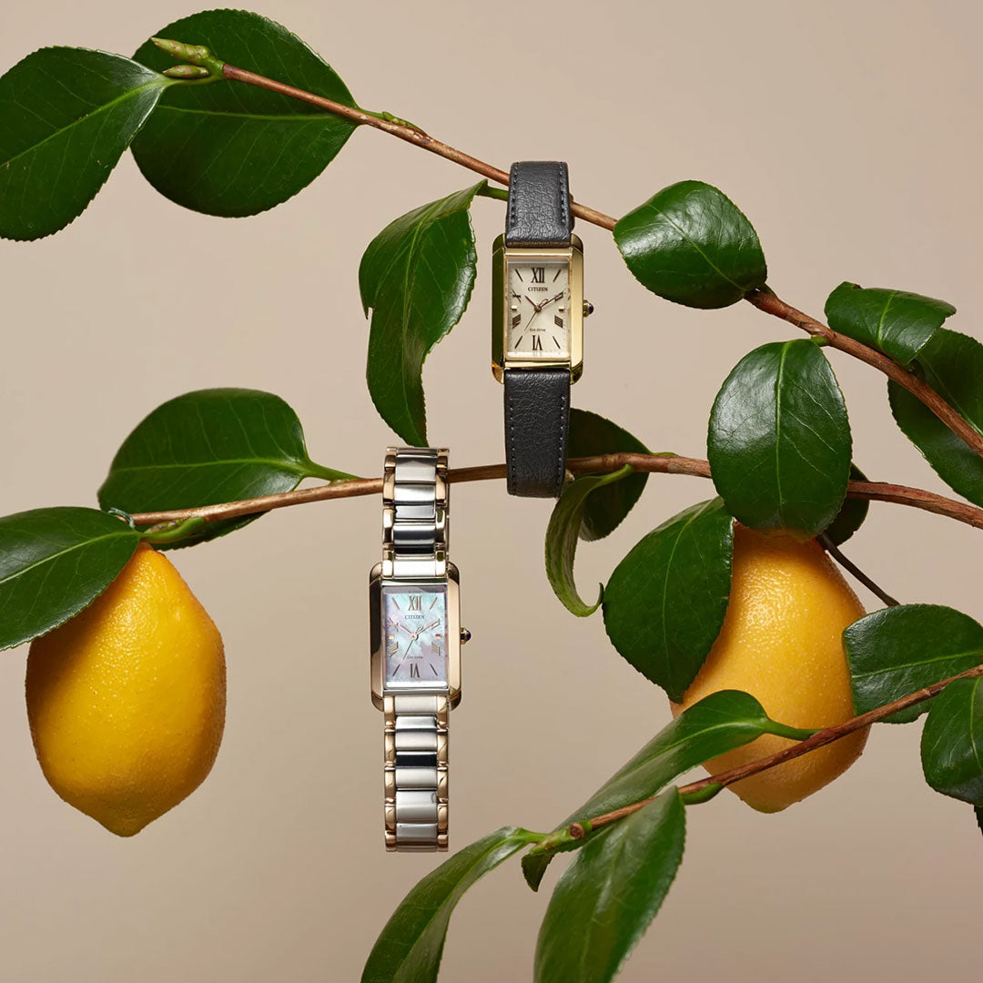 Two CITIZEN wristwatches hang on a leafy branch with yellow lemons: the top has a black leather strap, while the bottom is the CITIZEN L Bianca Eco-Drive 19.5mm Watch with a metal band. The background is plain light beige.