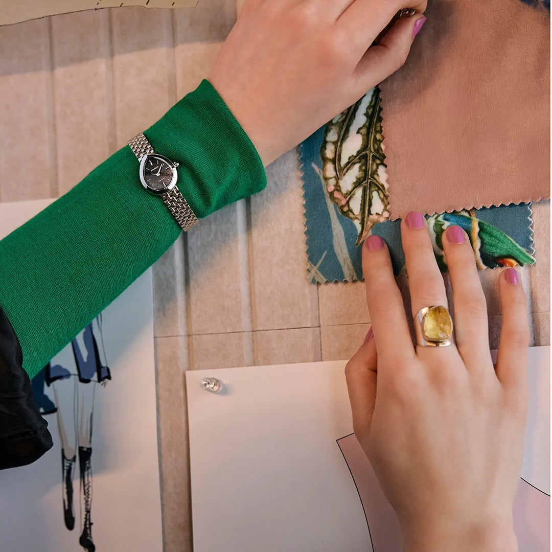 Two hands with painted nails touch fabric swatches on a wall; one wrist wears the CITIZEN L Rainell Eco-Drive 20mm Watch by CITIZEN, and one hand displays a large yellow gemstone ring.