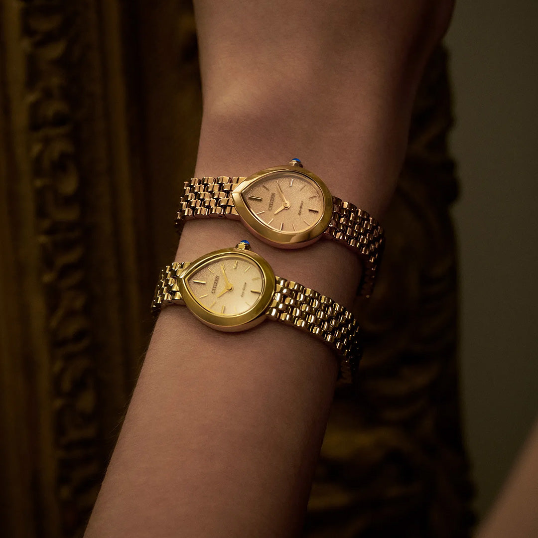 A close-up of a wrist adorned with two CITIZEN L Rainell Eco-Drive 20mm watches, each by CITIZEN, showcasing teardrop-shaped faces and elegant yellow gold-tone stainless steel bands against an ornate gold backdrop—emphasizing the refined minimalism and sophistication of the CITIZEN L Rainell collection.
