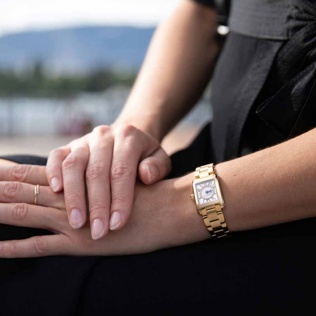 With hands resting gently on their lap, a person wears the Frederique Constant Classics Carree Ladies Quartz 30mm x 21mm Watch. Its elegant polished gold case and Roman numeral indexes stand out beautifully. The blurred background hints at an outdoor setting with water and greenery, while the person's black attire accentuates the watch's sophisticated allure.