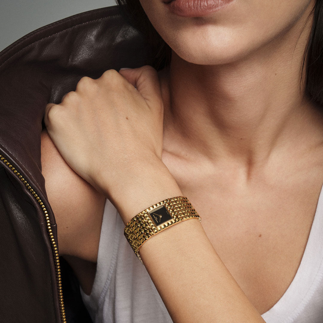 A person wearing a white tank top and brown jacket rests their hand on their shoulder, revealing the Frederique Constant Classics Manchette Quartz 25.7mm watch—its refined design and subtle Clou de Paris-inspired details adding an elegant touch of Frederique Constant sophistication to the look.