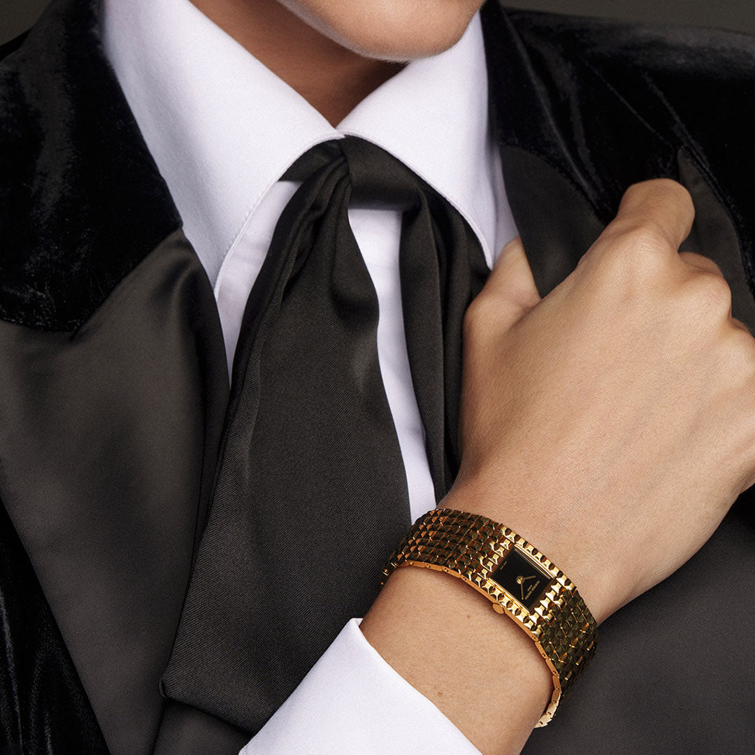 A person in a black velvet suit, white shirt, and black satin tie holds their suit lapel. On their wrist is the Frederique Constant Classics Manchette Quartz 25.7mm Watch, distinguished by its patterned gold bracelet and refined Clou de Paris motif—a subtle touch from Frederique Constant’s Classics Manchette collection.