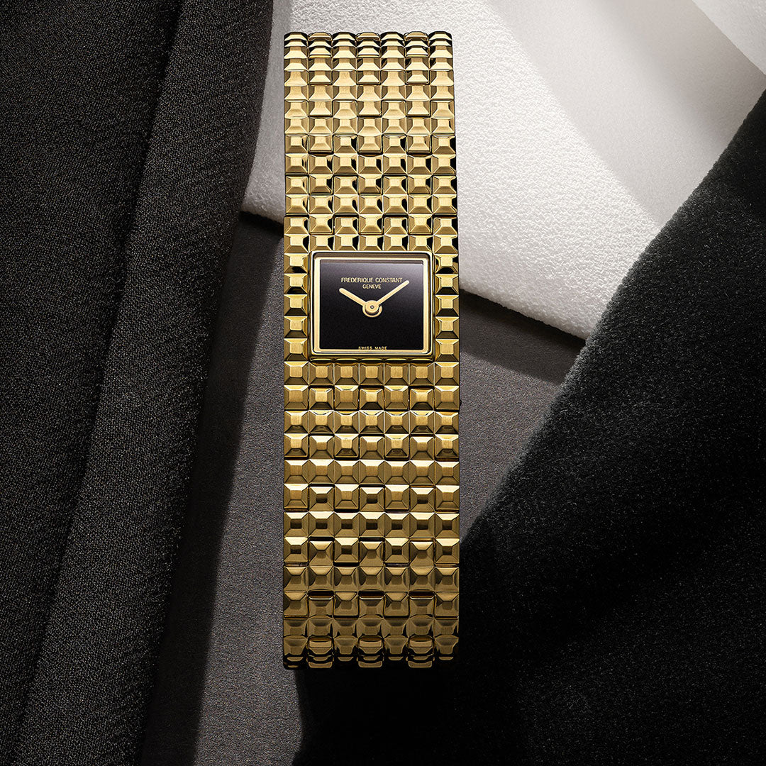The Frederique Constant Classics Manchette Quartz 25.7mm Watch, featuring a square gold case and a Clou de Paris textured bracelet, is displayed on an arrangement of black, gray, and white fabrics.