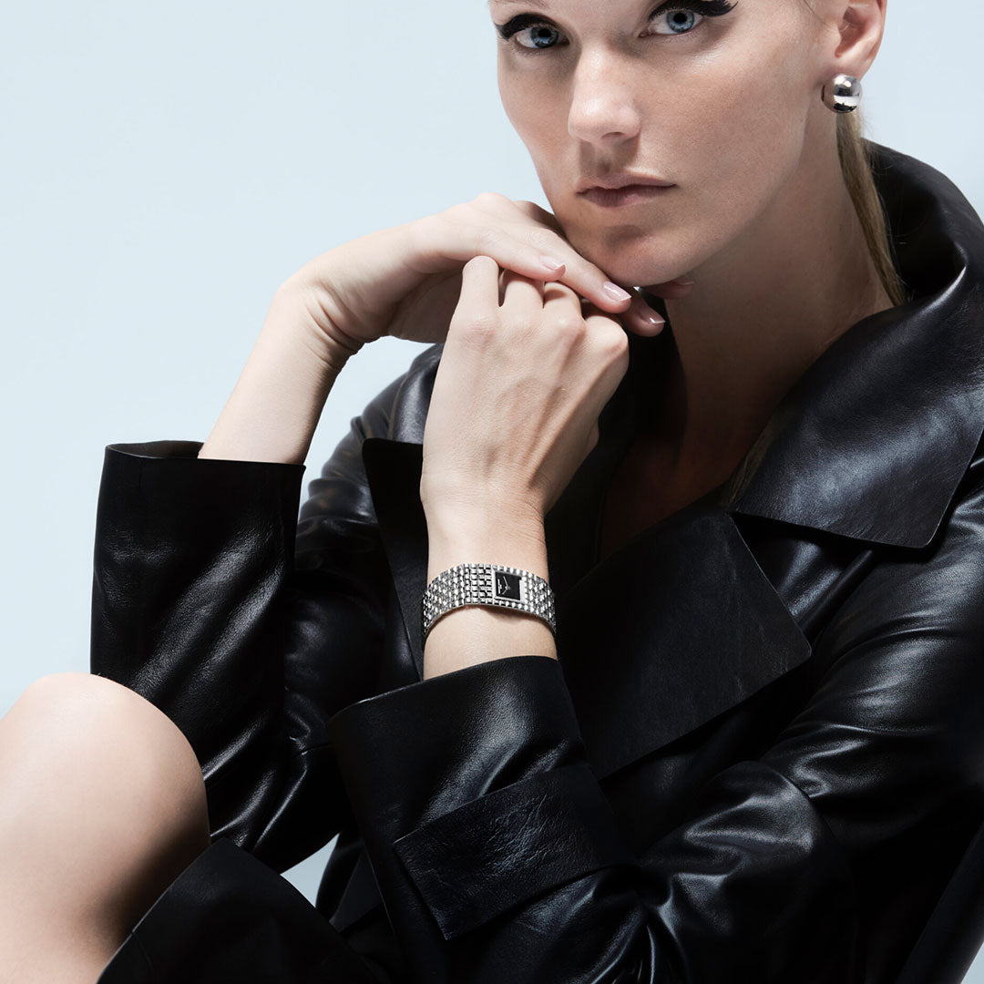 A woman in a black leather coat crosses her arms, displaying a Frederique Constant Classics Manchette Quartz 25.7mm Watch and silver earrings. She has light skin and an intense gaze against a plain light background.